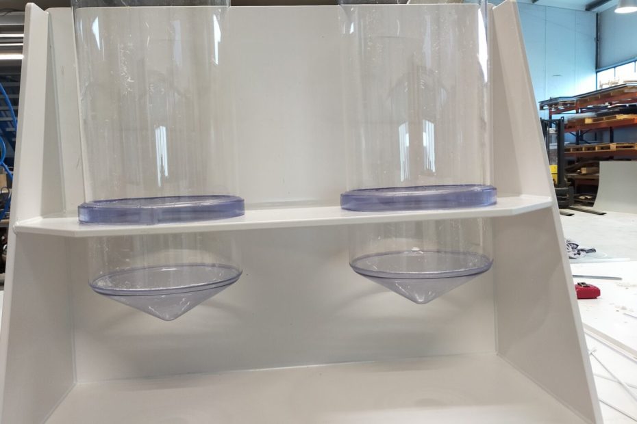 Vitrine plexiglas sur mesure en fabrication, avec deux cuvettes cylindriques hautes et deux supports inclinés translucides sur une étagère blanche.
