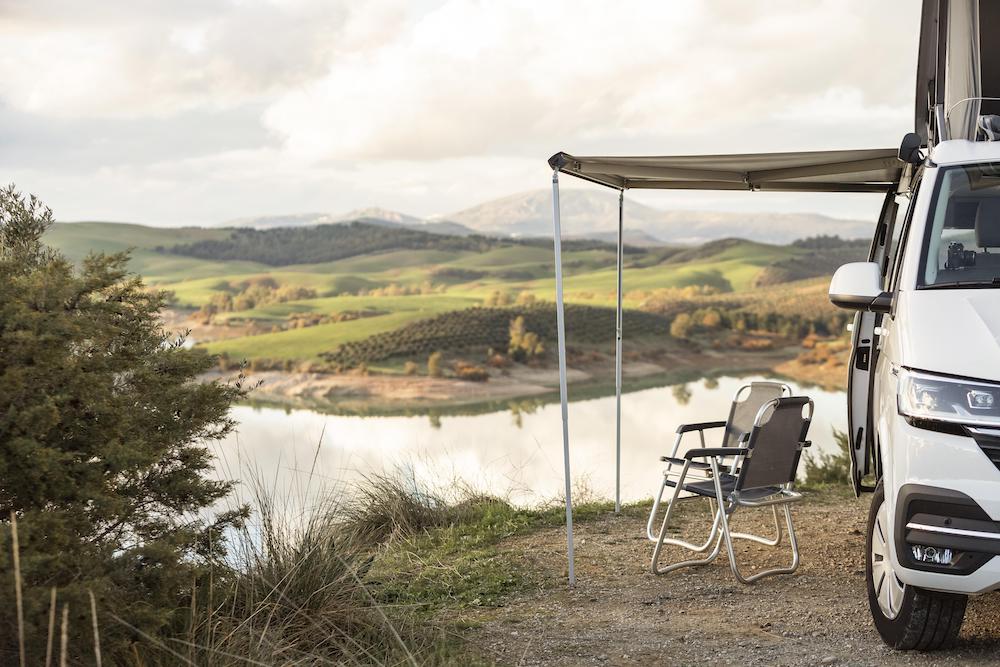 Vue d'un van aménagé près d'un lac, avec une pergola et une chaise pliante au premier plan, prêt pour un moment de repos en pleine nature. Incorporation naturelle de « fabriquer douche van personnalisée » si pertinent.