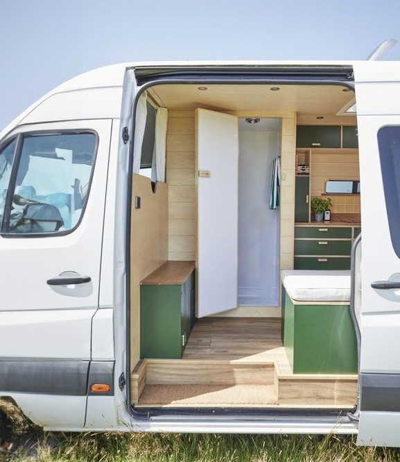 Intérieur d’un van aménagé avec douche et kitchenette, porte ouverte sur coin douche et rangements, sur plancher bois. fabrication douche van aménagé intégrée.